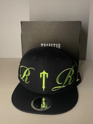 Trapstar keps - Säljer en svart fitted keps från Trapstar med stor limegrön broderad text runt hela kepsen. Klassisk platt skärm. Materialet är tåligt och kepsen har en modern streetwear-look som sticker ut. Storlek 7 1/4 = 58cm