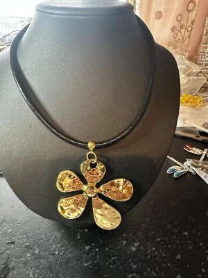 Halsband med stor guldfärgad blomma - Unikt halsband med ett stort, hamrat blomformat hänge i guldfärgad metall. Hänget sitter på en läderrem ger en cool kontrast till det glänsande guldet. Perfekt statement-piece för dig som gillar att sticka ut med accessoarer.