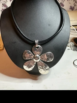 Silvrigt halsband med stor blomma - Unikt halsband med ett stort, hamrat blomformat hänge i silverfärgad metall. Hänget har fem breda kronblad och sitter på ett svart, läderrem detta är ca 47 cm långt. Perfekt för dig som vill sticka ut med ett statement-smycke och gillar retrovibbar.