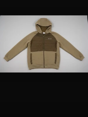 Beige pufferjacka från Columbia - Snygg beige vår jacka från Columbia med bruna detaljer och huva. Jackan har dragkedja framtill, två fickor och quiltad insida för extra värme. Perfekt för kalla dagar och har en modern, avslappnad look. Materialet är en mix av syntet och vaddering.