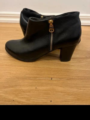 Svarta stövletter med klack och dragkedja - Säljer ett par svarta stövletter med snygg klack och guldfärgad dragkedja på sidan. Skorna har rund tå och är tillverkade i skinn, vilket ger en stilren och klassisk look. Perfekta för dig som gillar enkel men cool design.
