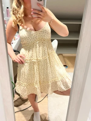 Blommig ljus kortklänning med volang - Söt ljus kortklänning med små blommor och volangdetaljer. Klänningen har smala axelband, v-ringning och en luftig passform som ger en romantisk vibe. Perfekt för varma dagar och sommarhäng.