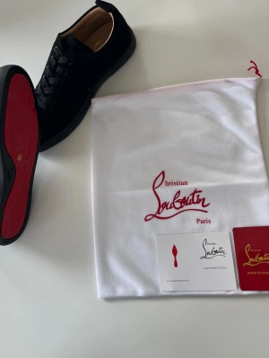 Svarta sneakers från Christian Louboutin - Snygga svarta sneakers från Christian Louboutin med klassisk röd sula och svarta skosnören. Skorna har en stilren design i läder och kommer med originalpåse, box och certifikat. Perfekta för dig som vill ha något exklusivt och trendigt.