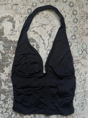 Svart halterneck topp - Snygg svart halterneck topp med djup urringning och breda band som man har runt nacken. Toppens material känns stretchigt och mjukt, perfekt för att skapa en cool och enkel look. Passar till många olika stilar och är lätt att matcha med andra plagg.