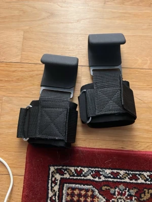 Svarta lyftkrokar för gymträning - Ett par svarta lyftkrokar med breda kardborreband och vadderade handlovsstöd. Perfekta för tunga lyft och styrketräning, ger extra grepp och stöd vid exempelvis marklyft. Tillverkade i slitstarkt material med metallkrok och nylonband.