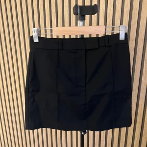 Svart kostymkjol med H&M - Stilren svart kortkjol med bälteshällor och diskret ficka bak. Klassisk rak modell som passar till många olika looks. Kjolen har en enkel design utan synliga detaljer framtill och är lätt att matcha.