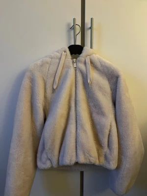 Beige teddyjacka med huva - Mysig beige teddyjacka med dragkedja framtill och stor huva. Jackan har dragsko i huvan och är gjord i supermjukt, fluffigt material som ger en avslappnad och trendig look. Perfekt för kyliga dagar när du vill vara både varm och snygg.