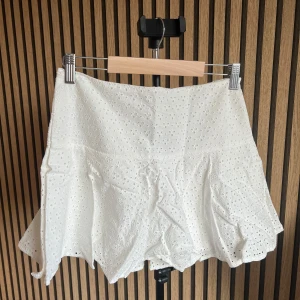 Vit kjol från H&M - Superfin vit kort kjol med broderade hålmönster och lätt utsvängd form. Kjolen har en feminin och luftig känsla, perfekt för varma dagar. Passar snyggt till både enkla toppar och stickade tröjor.