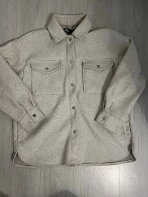 Beige overshirt med fickor - Stilren beige overshirt från Vila med klassisk krage och två stora bröstfickor med lock och knapp. Overshirten har tryckknappar framtill och vid ärmsluten, samt en rak och något längre passform. Perfekt lager på lager-plagg för en avslappnad look.