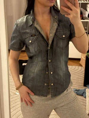 Lee Kortärmad jeansskjorta med tryckknappar - Säljer en snygg Populär Lee mörkblå jeansskjorta med korta ärmar och tryckknappar framtill. Skjortan har två bröstfickor med lock och detaljerade sömmar för en cool look. Perfekt till jeans eller kjol för en avslappnad stil. Passar även S