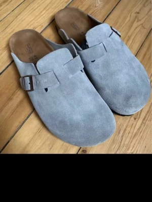 Birkenstock boston tofflor - Säljer ett par stilrena gråa tofflor från Birkenstock i mjuk mocka. De har en bred rem med metallspänne och klassisk korksula för extra komfort. Perfekta för dig som vill ha både snygg och avslappnad stil hemma eller ute.