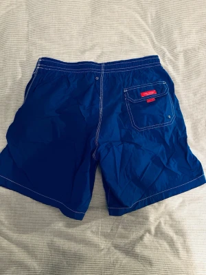 Blå badbyxor från tino cosma  - Snygga blå badbyxor från tino cosma med vit dragsko i midjan och kontrasterande vita sömmar. Bakficka med röd logotypetikett och knappstängning. Klassisk modell i snabbtorkande material, perfekt för stranden eller poolen.