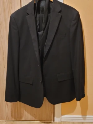 Svart kostym från H&M - Klassisk svart kostym från H&M med tvåknäppt kavaj och matchande väst. Kavajen har smala slag, två framfickor med lock och innerficka. Fodrad insida för extra komfort. Perfekt för dig som vill ha en stilren och enkel look.