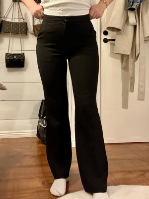 Svarta utsvängda  kostymbyxor - Svarta byxor med utsvängda ben och hög midja. Byxorna har en stilren och enkel design utan synliga fickor eller detaljer. Perfekta för dig som gillar en klassisk och tidlös look. Materialet ser ut att vara stretchigt och bekvämt.