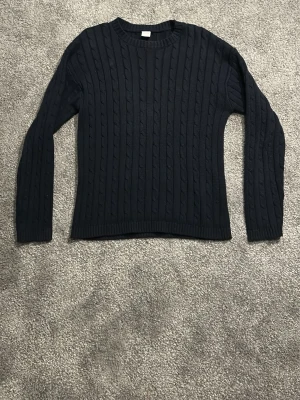 Mörkblå kabelstickad tröja - Kabelstickad tröja i mörkblått med rund halsringning och långa ärmar. Tröjan har klassiskt flätmönster och ribbade muddar vid ärmslut och nederkant. Perfekt för kyliga dagar och enkel att matcha med jeans eller kjol.