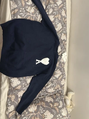 Mörkblå stickad tröja från Ami Paris - Mörkblå stickad tröja från Ami Paris med vitt hjärta och bokstaven A broderat på bröstet. Tröjan har rund halsringning, långa ärmar och breda ribbade muddar vid ärmslut och nederkant. Perfekt för en stilren och trendig look.