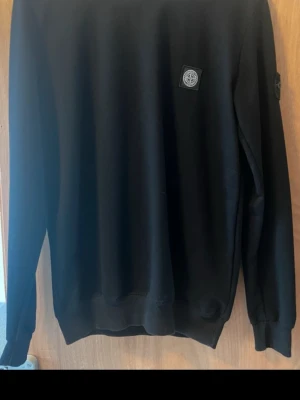 Svart sweatshirt från Stone Island - Svart sweatshirt från Stone Island med klassisk logotyp-patch på vänster ärm och en mindre patch på bröstet. Tröjan har rund halsringning, ribbade muddar och är tillverkad i mjukt material. Perfekt för en stilren och avslappnad look.