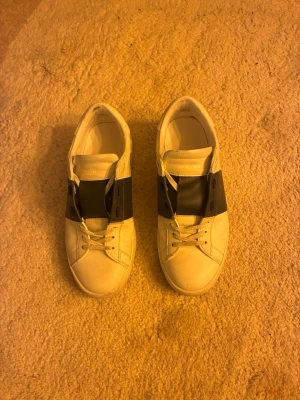 Vita sneakers med svart detalj - Snygga vita sneakers med bred svart rem över vristen och klassisk snörning. Skorna har en stilren och minimalistisk design med rund tå och platt sula. Tillverkade i läderliknande material och passar perfekt till streetwear eller casual outfits.