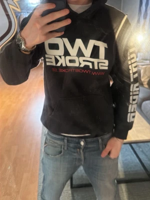 Svart hoodie TWO STROKE - Cool svart hoodie från TWO STROKE med stor vit och röd logga på bröstet och tryck på ärmen. Baksidan har ett färgglatt motiv med stunt rider på motorcykel. Klassisk huva och känguruficka framtill. Perfekt för dig som gillar motorcyklar och streetstyle.