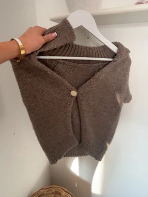 Brun stickad väst med knapp - Mysig brun stickad väst med en dekorativ knapp framtill. Västen har en djup V-ringning och ribbstickade kanter. Perfekt att bära över en skjorta eller t-shirt för en trendig lager-på-lager-look. Från Zara i xs