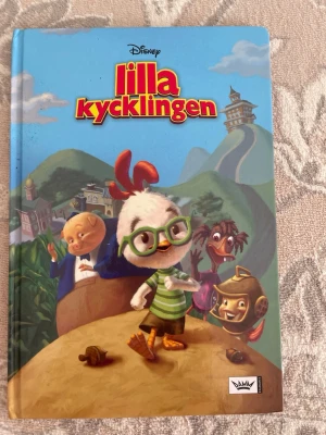 Lilla Kycklingen bok Disney - En färgglad barnbok med hårda pärmar från Disney, 'Lilla Kycklingen'. Omslaget har tecknade figurer i glada färger och boken är fylld med illustrationer och text. Perfekt för dig som gillar roliga och äventyrliga berättelser.