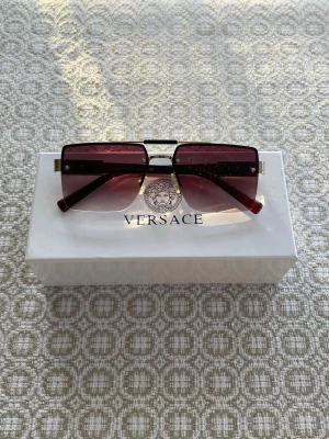 Rimless Vinted Versace solglasögon - Snygga solglasögon från Versace med rektangulära, vinröda glas och metallbågar. Kommer med originaletui och box. Perfekt accessoar för dig som vill sticka ut med en exklusiv look. En eldre utgåva från Versace men skicket är fortfarande bra.