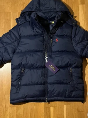 Mörkblå pufferjacka från Polo Ralph Lauren - Snygg mörkblå pufferjacka från Polo Ralph Lauren med huva och dragkedja framtill. Jackan har två sidofickor med dragkedja och den klassiska röda loggan broderad på bröstet. Perfekt för kalla vinterdagar och riktigt trendig look.