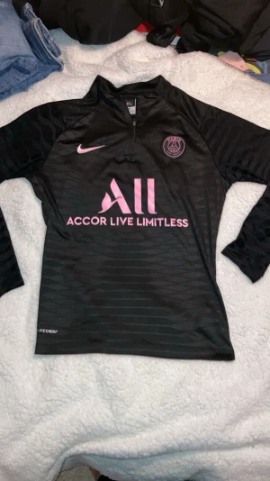 Svart PSG träningströja från Nike - Svart långärmad träningströja från Nike med PSG-logga och rosa detaljer. Tröjan har halv dragkedja framtill, Dri-FIT-material och texten 'Accor Live Limitless' på bröstet. Perfekt för fotbollsträning eller gymmet.