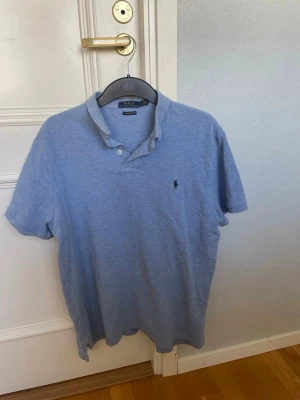 Blå pikétröja från Polo Ralph Lauren - Klassisk ljusblå pikétröja från Polo Ralph Lauren med korta ärmar och krage. Tröjan har två knappar vid halsen och den ikoniska loggan broderad på bröstet. Perfekt till jeans eller shorts för en avslappnad stil.