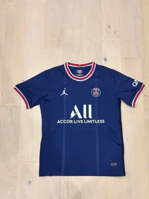 PSG matchtröja blå Jordan x Nike - Snygg blå Paris Saint-Germain matchtröja från Nike och Jordan, med vita och röda detaljer på krage och ärmslut. PSG-logga, Jordan-logga och sponsortryck på bröstet. Tillverkad i lätt och ventilerande funktionsmaterial, perfekt för fotbollsträning.