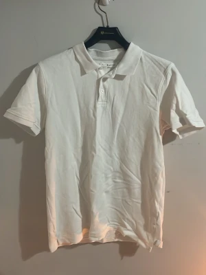 Vit pikétröja från S.C.W - Klassisk vit pikétröja från S.C.W med krage och två knappar framtill. Tröjan har korta ärmar och är tillverkad i ett mjukt bomullsmaterial. Enkel och stilren design som passar till det mesta.