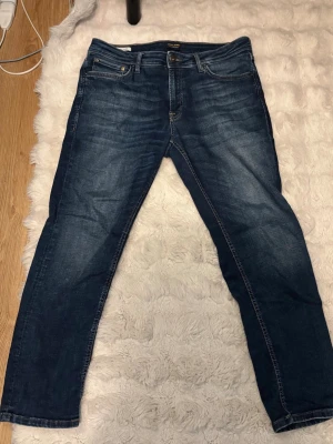 Mörkblå jeans från Jack & Jones - Säljer ett par klassiska mörkblå jeans från Jack & Jones med fem fickor och snygga slitningar framtill. Jeansen har normal passform och är tillverkade i ett robust denimtyg. Perfekta för en avslappnad och stilren look. Modellen är Clark. 33x30
