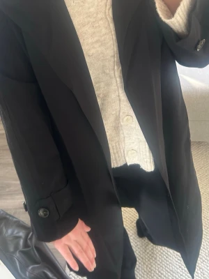 Svart trenchcoat från Gina Tricot - Stilren svart trenchcoat från Gina Tricot med klassisk krage, knappar framtill och bälte i midjan. Långa ärmar med knappdetaljer vid ärmslut och slits baktill☀️