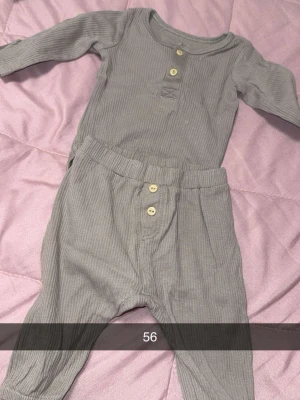 Grå ribbat babyset med knappar - Säljer ett gulligt tvådelat babyset i ribbat grått tyg. Setet består av en långärmad body med rund hals och tre träfärgade knappar framtill samt matchande byxor med resår i midjan och dekorativa knappar. Perfekt för mysiga dagar hemma.