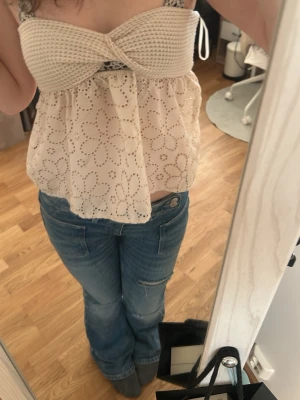 Beige broderad linnetopp - Söt beige linnetopp med virkad överdel och broderade blommor på den luftiga nederdelen. Toppen har axelband och en twistad detalj framtill. Perfekt för varma dagar och ger en bohemisk känsla.