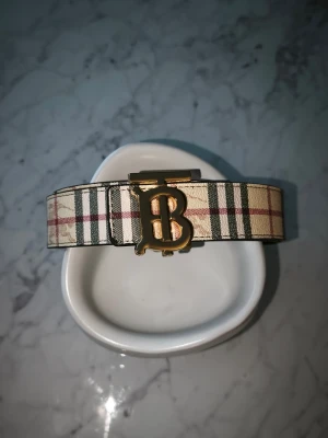 Burberry bälte med guldigt märke - Tvär fett bälte från Burberry, väldigt enkel att styla och har en unik "funktion", man kan vända bältet ut o in och byta sida på loggan. Det gör att bältet då blir svart och på bild tre så kan du se hur det kommer att se ut (loggan blir också på den sidan)  