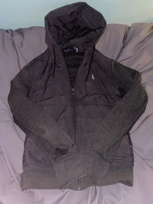 Mörkgrå hoodiejacka från Polo Ralph Lauren - Snygg mörkgrå hoodiejacka från Polo Ralph Lauren med dragkedja framtill och broderad logga på bröstet. Jackan har quiltade partier, långa ärmar i mjukt material och praktisk huva med snörning. Perfekt för lager på lager under kyliga dagar. Som man ser så har båda ärmarna hål i sig, därav sitt låga pris. PRIS KAN ALLTID DUSKUTERAS!!