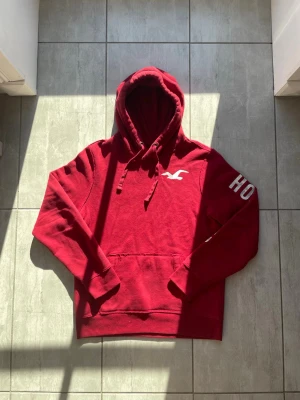 Röd hoodie från Hollister - Snygg röd hoodie från Hollister med stor huva och dragsko. Klassisk logga i vitt på bröstet och texten 'HOLLISTER' tryckt på ena ärmen. Mjuk bomullsmix och känguruficka framtill. Perfekt för en avslappnad och trendig look. Vid frågor eller funderingar skriv gärna!