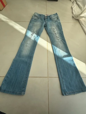 BOOTCUT Blå bootcut jeans från Måndag - Snygga ljusblå bootcut jeans från Måndag med slitningar och slitna detaljer på låren. Jeansen har klassisk femficksmodell, dubbla knappar i midjan och cool tvättad look. Perfekta för dig som gillar retrovibbar och avslappnad stil.
