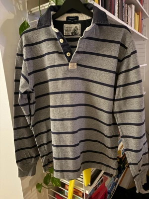 Randig tröja från Gant - Klassisk tröja från Gant i grått med mörkblå horisontella ränder. Tröjan har en mörkblå krage, knappar vid halsen. Använd en gång!