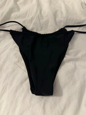 Svart bikiniunderdel med snörning - Säljer en svart bikiniunderdel med tunna snören i sidorna. Klassisk och enkel design som passar perfekt till sommaren. Materialet känns mjukt och följsamt mot huden.