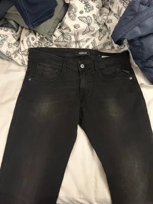 Svarta Replay jeans med smal passform - Säljer ett par svarta jeans från Replay med smal passform och klassisk femficksdesign. Jeansen har subtila slitningar framtill och snygga detaljer på bakfickorna. Tillverkade i stretchigt material för extra komfort.