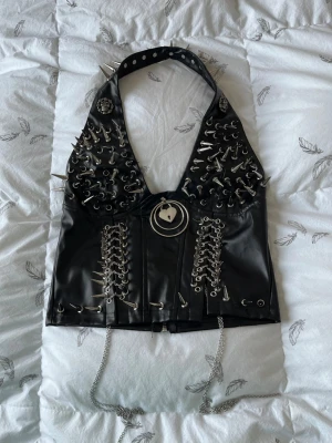 Svart korsettopp med nitar och kedjor - Unik svart korsettopp i syntetmaterial med massor av silverfärgade nitar, spikes och kedjor. Toppen har snörning framtill, stora metallringar och ett dekorativt hänglås i mitten. Perfekt för dig som gillar alternativ stil och vill sticka ut.