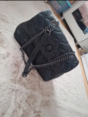 Svart quiltad handväska med kedja - Snygg svart quiltad handväska med silverfärgad kedjerem och läderdetaljer. Väskan har ett diskret emblem på framsidan och dragkedjefack på baksidan. Perfekt för dig som gillar en stilren och trendig look.