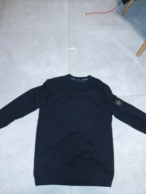 Svart sweatshirt från Stone Island - Säljer en svart sweatshirt från Stone Island med det klassiska kompassmärket på vänster ärm. Tröjan har rund hals, långa ärmar och ribbade muddar. Materialet är mjukt och bekvämt, perfekt för en avslappnad stil. Stort ton-i-ton broderi på bröstet.