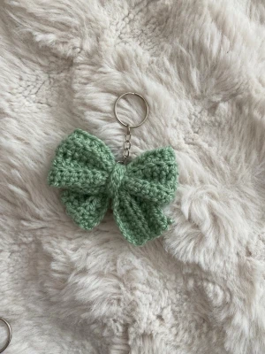 Stickad rosett nyckelring grön - Söt handgjord nyckelring med en stickad rosett i ljusgrönt garn. Rosetten är fäst på en metallkedja med nyckelring, perfekt att hänga på väskan eller nyckelknippan. Materialet är mjukt garn och metall.
