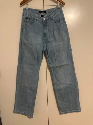 Ljusblå raka jeans från South Pole - Ljusblå jeans från South Pole med rak Bred passform och klassisk femficksdesign. Jeansen har hög midja, silverfärgade knappar och detaljerade sömmar. Tillverkade i denim med en avslappnad vintagekänsla. Perfekta för en casual och trendig look.