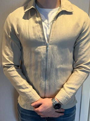 Beige tunn J.Lindeberg overshirt med dragkedja - Säljer en stilren beige jacka i tunnare material med klassisk krage och dragkedja framtill. Jackan har raka ärmar och enkel design. 