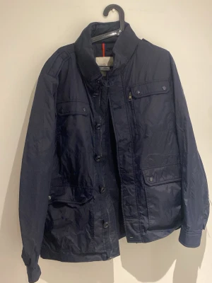 Mörkblå vindjacka från Moncler - Snygg mörkblå vindjacka från Moncler med hög krage och flera fickor framtill. Jackan har både dragkedja och knappar, samt diskret logga på kragen. Tillverkad i ett lätt, glansigt material som står emot vind. Perfekt för dig som vill ha en stilren och funktionell jacka.