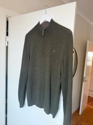 Olivgrön halvzip-tröja från Morris - Snygg olivgrön stickad tröja från Morris med halv dragkedja och liten broderad logga på bröstet. Tröjan har hög krage och ribbade muddar vid ärmslut och nederkant. Perfekt för lager på lager och stilren look. Large men sitter som M